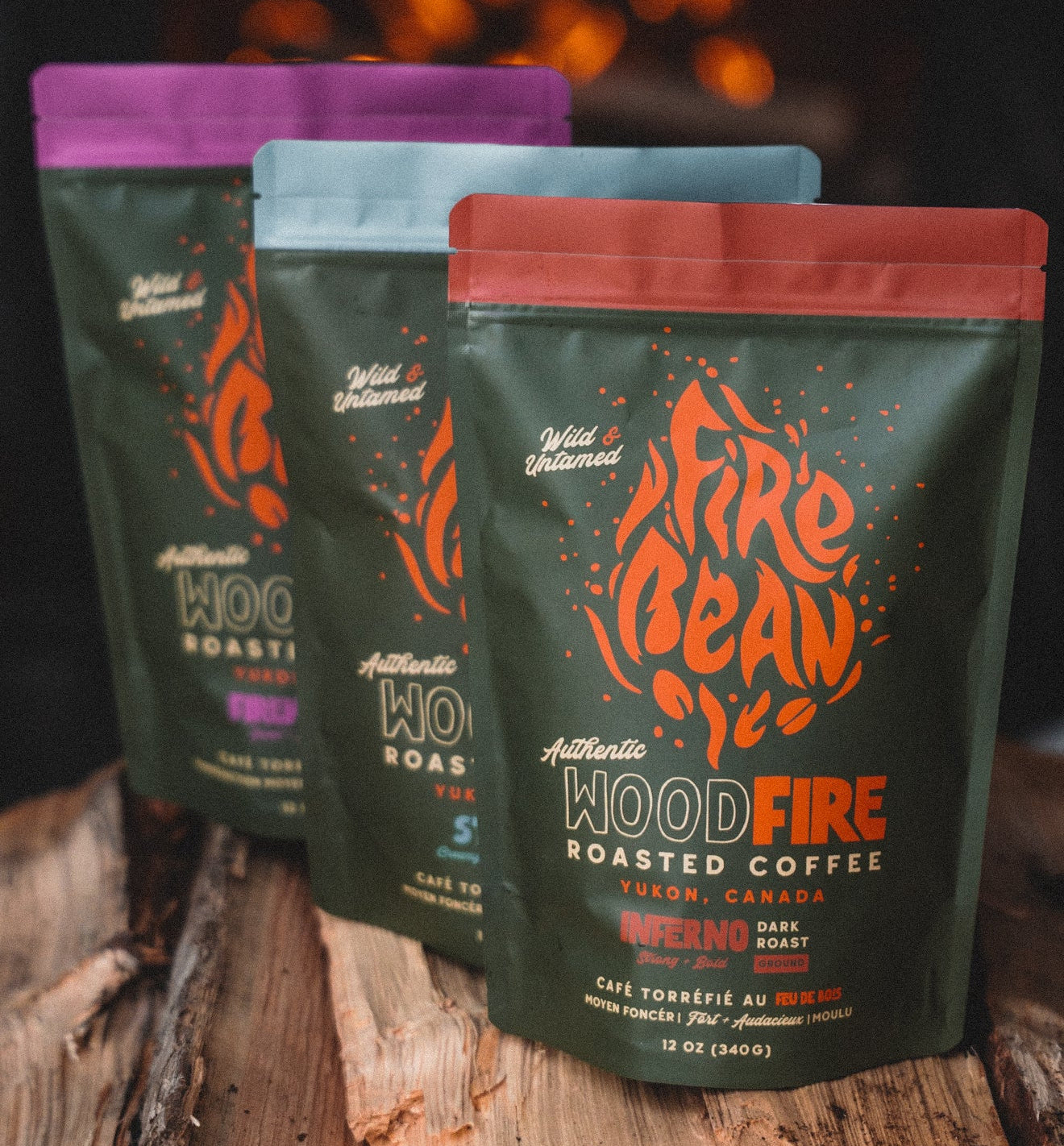 Three packages of Fire Bean WoodFire roasted coffee on wooden logs with a fireplace in the background.