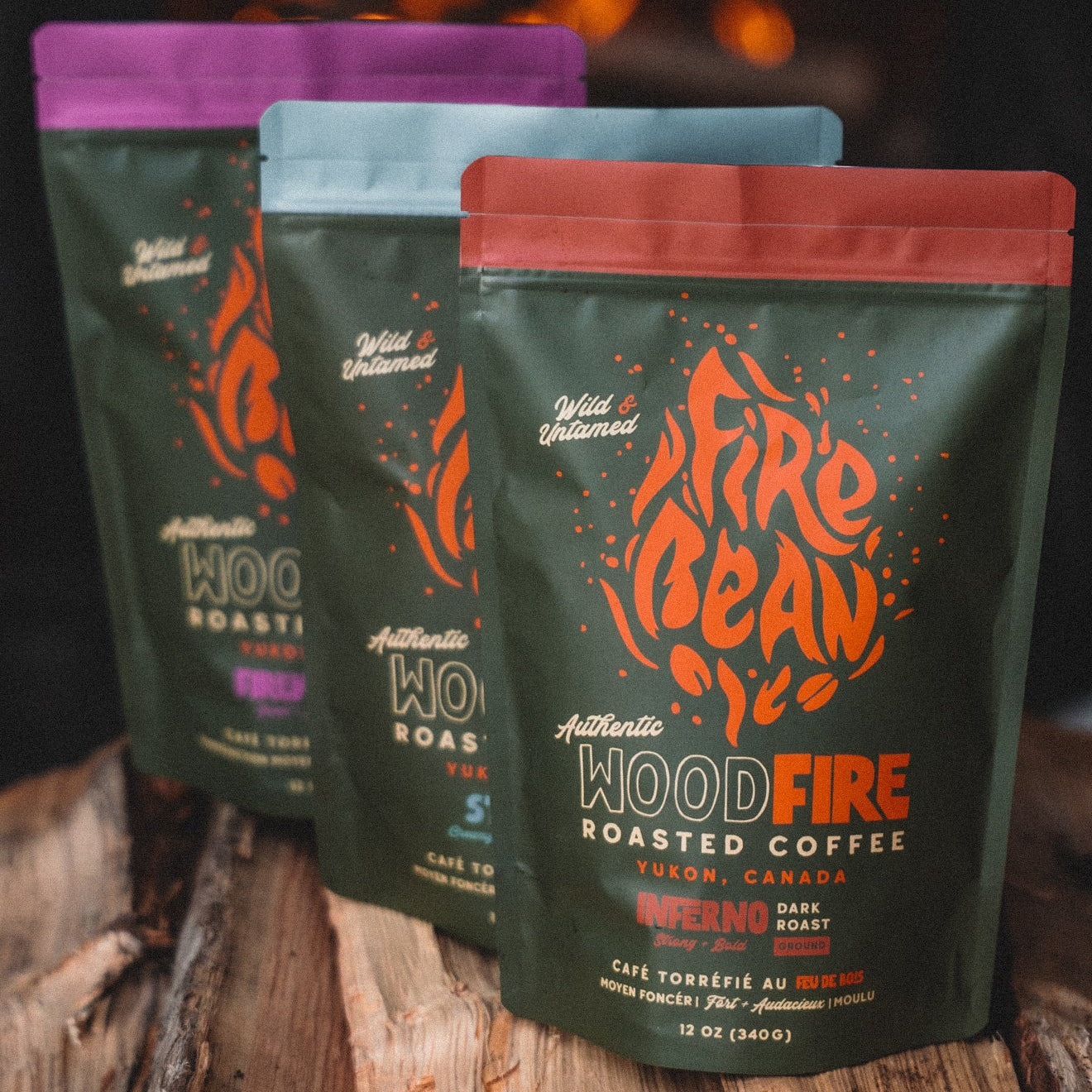 Three packages of Fire Bean WoodFire roasted coffee on wooden logs with a fireplace in the background.