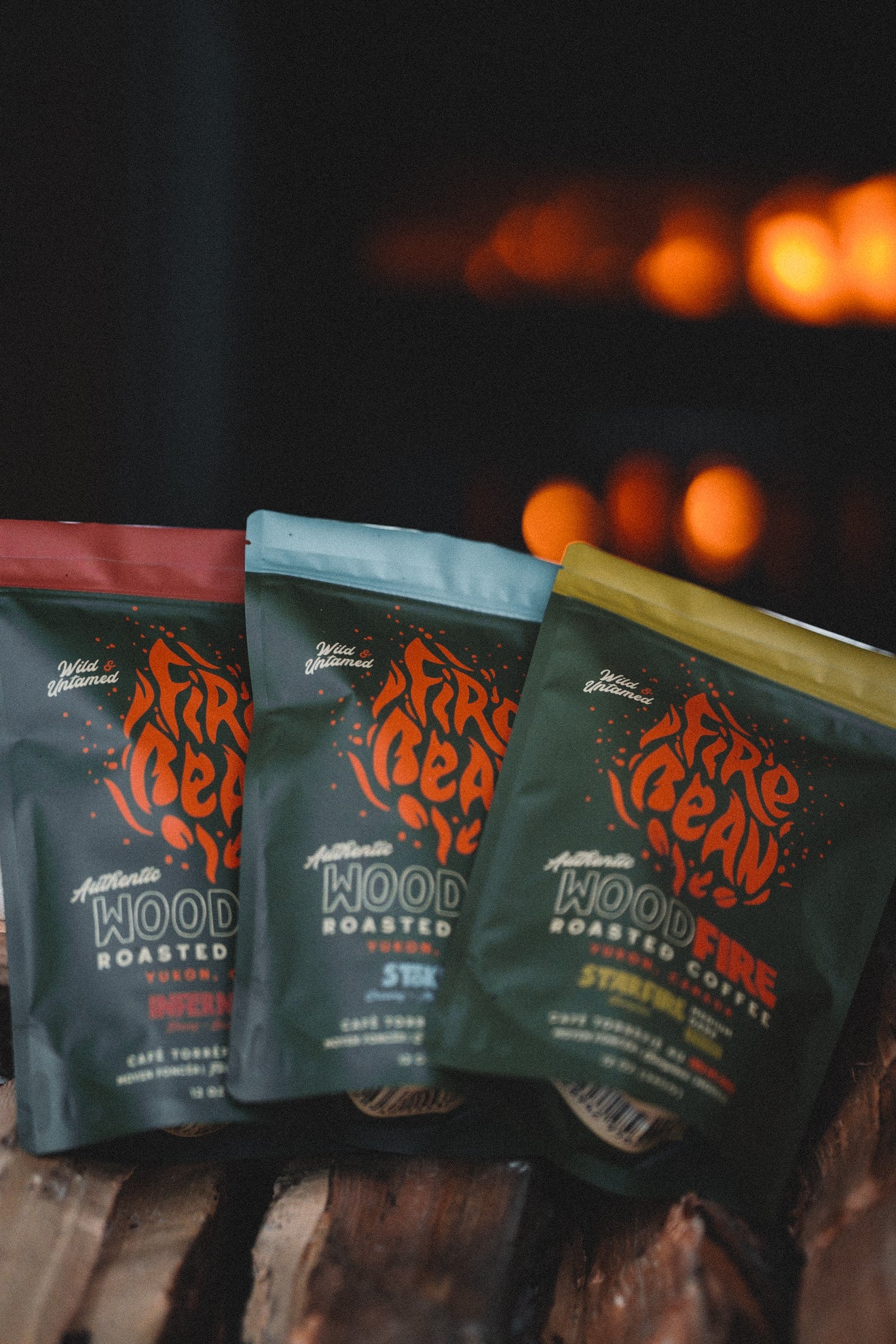 Three packages of FireBean coffee on a wooden surface with a fireplace in the background