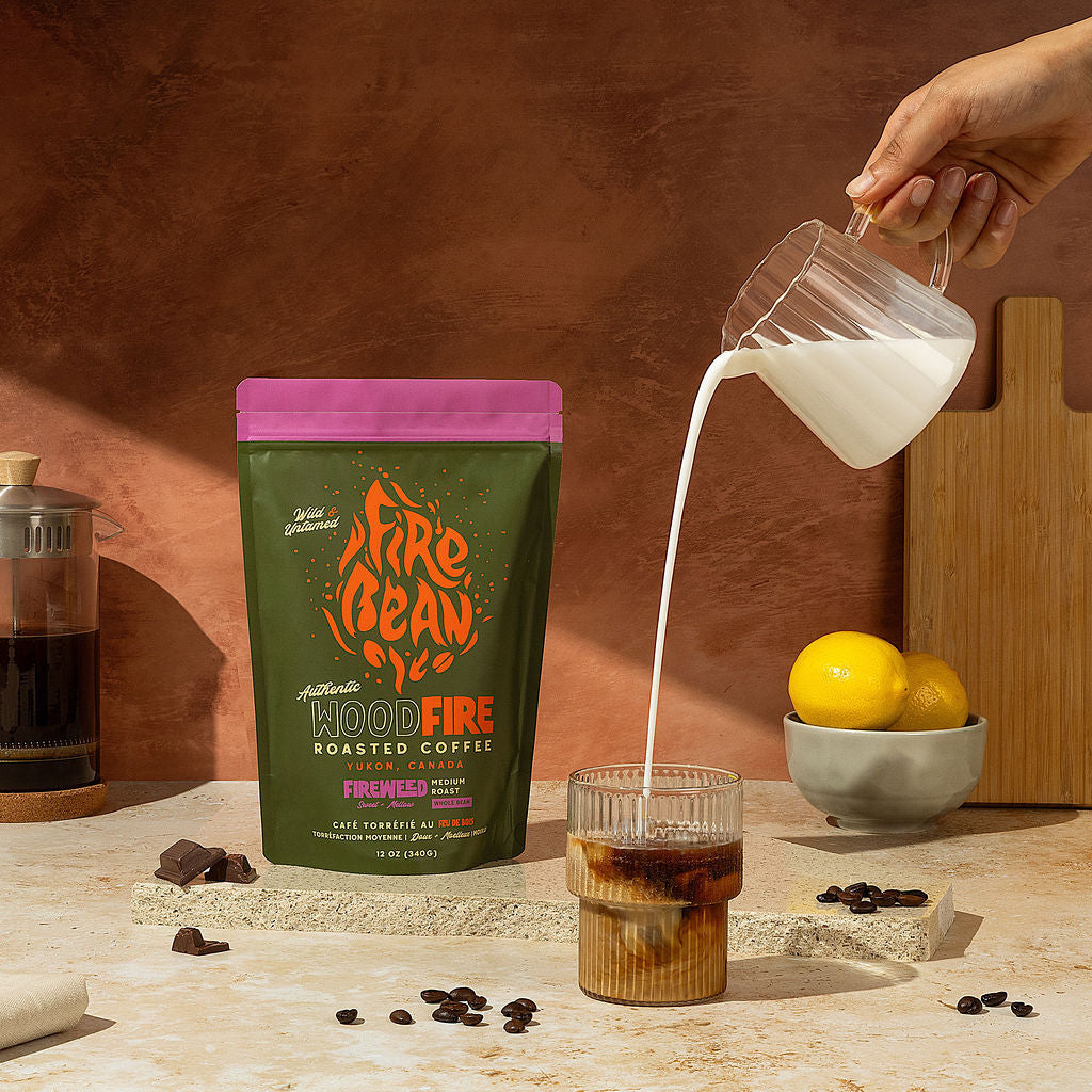 Person pouring milk into a glass of iced coffee next to a bag of Fire Bean Hood Fire coffee on a countertop.